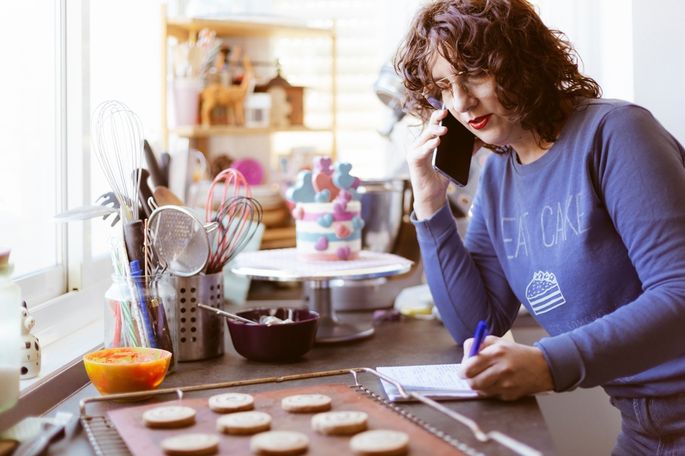 Mulher empreendedora faz uma anotação em um caderno enquanto fala ao telefone em uma cozinha onde prepara bolos e doces