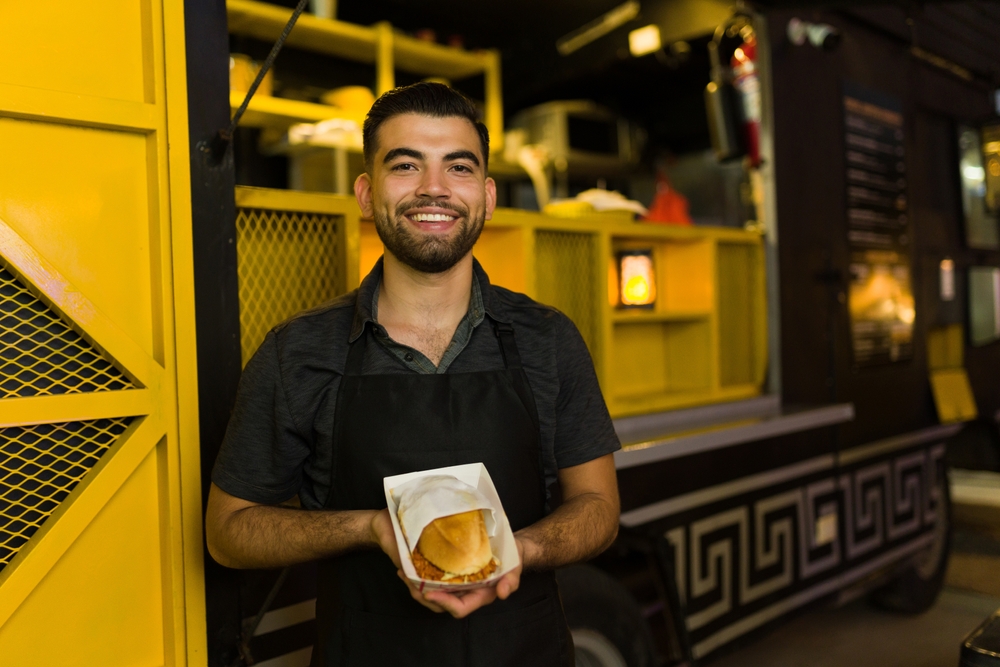 Homem empreendedor de avental sorri em frente a um food truck com um lanche embalado nas mãos