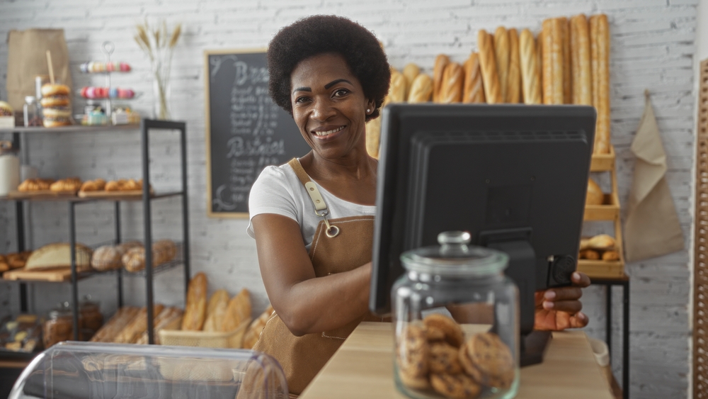 Mulher de avental, empreendedora, sorrindo, no caixa de uma padaria e confeitaria